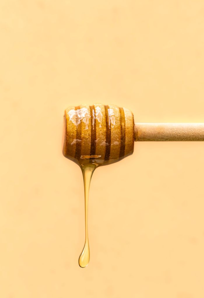 services-01 Close-up of honey drizzling from a wooden dipper against a soft yellow background.
