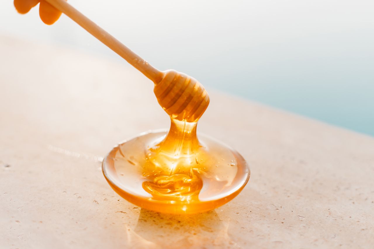about-bg Golden honey flowing from a wooden dipper onto a glass plate, captured in warm daylight.